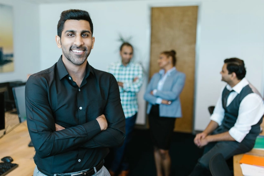A confident young professional standing in a modern office with colleagues interacting in the background