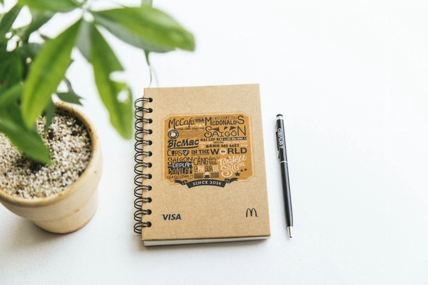 A spiral notebook and pen on a white background next to a potted plant, creating a minimalistic workspace vibe